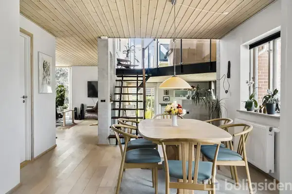 Et rundt spisebord med seks stole står i en lys stue. En trappe fører op til en åben balkon. Store vinduer og planter skaber en indbydende atmosfære.