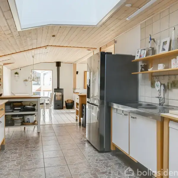 Et moderne køkken med hvide skabe og en stor metal-køleskab. Rummet er lyst med et træloft og hylder med dekorationer. I baggrunden ses en spiseplads og en brændeovn.