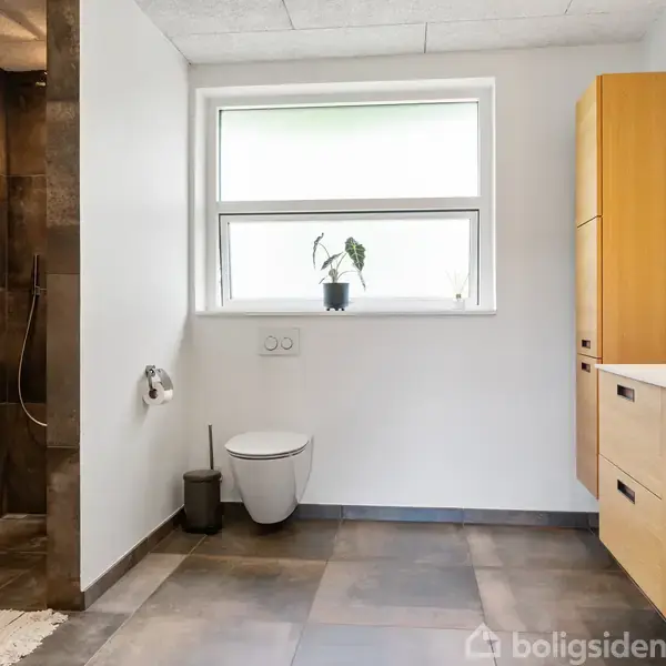 Badeværelse med et dusjområde til venstre, toilet og plante ved et vindue i midten, samt et træskab med vask til højre. Lyse vægge og gulvfliser.