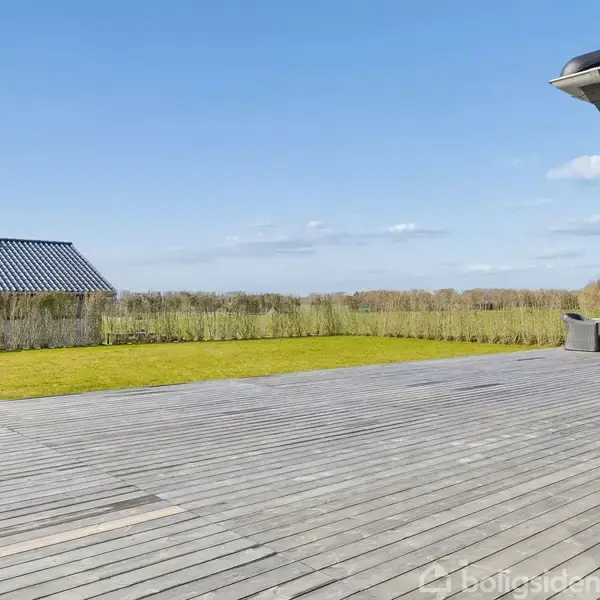 En stor træterrasse strækker sig ud fra et moderne hus, omgivet af en velholdt græsplæne og med udsigt til en mark under en klar blå himmel.