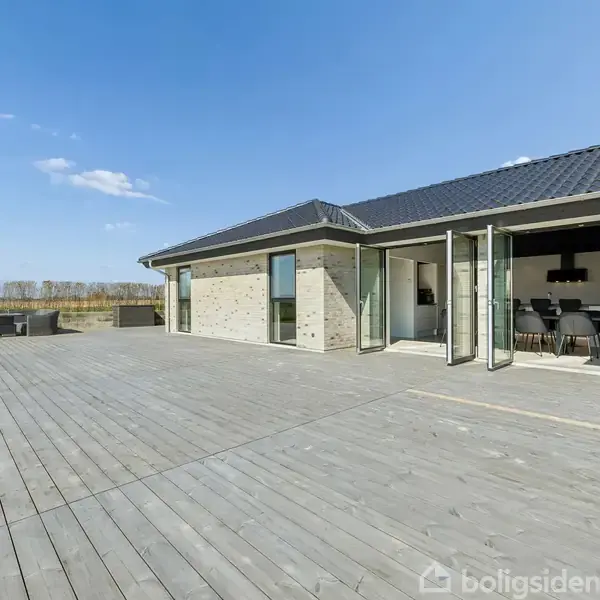 Et moderne hus med store glasdøre fører ud til en stor træterrasse. Omgivet af en grøn have, under en klar blå himmel.