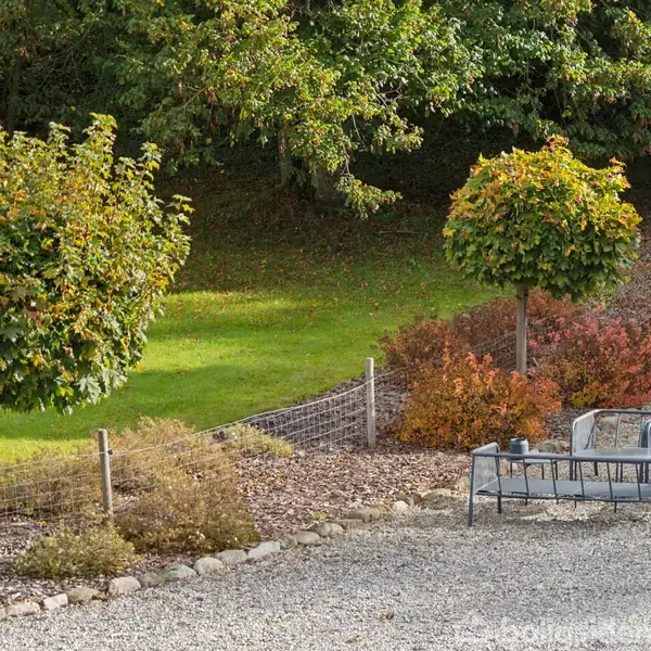Træer står stille i en have ved siden af et grusområde. En siddegruppe med havemøbler er placeret tæt på, omgivet af buske og grøn plæne.