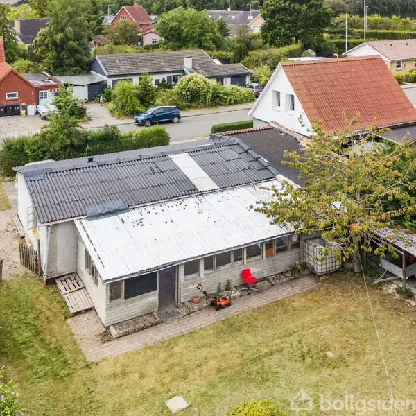Et ældre hus med en blanding af hvide og grå tage står i en forstad. En rød stol er placeret foran huset, omgivet af en have med træer og buske.