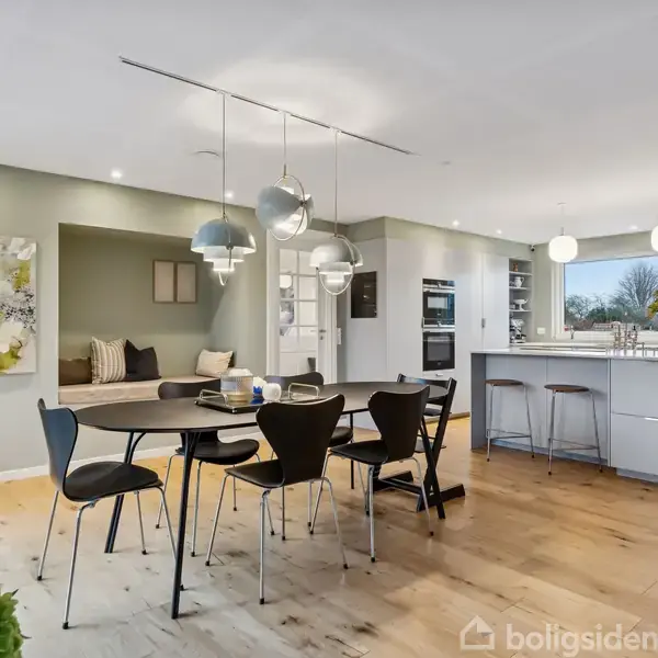 Et moderne køkken-alrum med et rundt spisebord og sorte stole. I baggrunden er en køkkenø med barstole. Rummet har trægulv, kunst på væggen og moderne belysning.