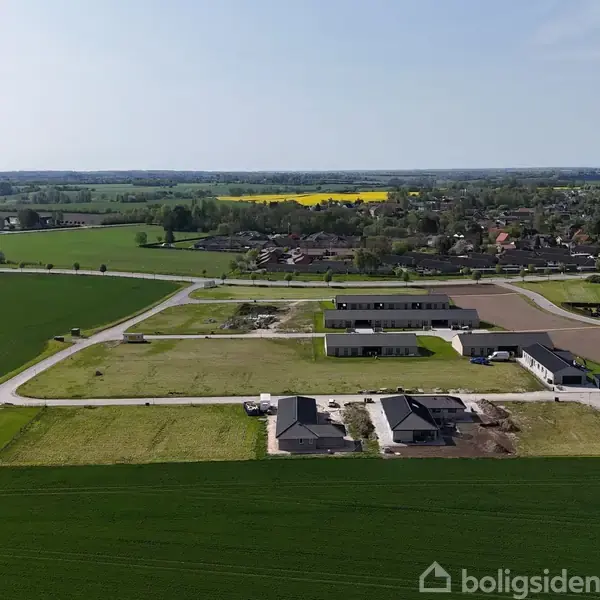 Luftfoto af nybyggeri med flere huse placeret i et åbent landskab omgivet af marker og en nærliggende landsby. Bygningerne ligger langs små veje i et parcelhuskvarter.