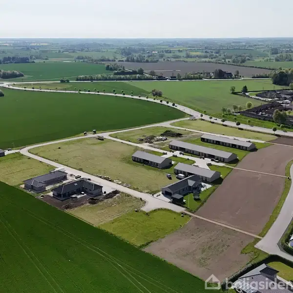 Boligområde set fra luften. Flere moderne huse står spredt på store parceller. Omgivet af åbne marker og landeveje, med en landsby i baggrunden.
