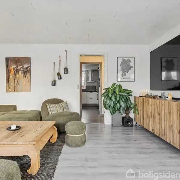 Grøn sofa står i en lys stue med gråt gulv. Ved siden af er et træbord, og overfor hænger et stort tv på væggen. En plante og billeder dekorerer rummet.