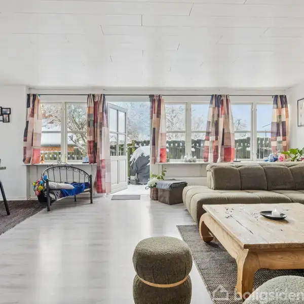 En stue med en stor, grøn sectional sofa, et træ sofabord og en lænestol. Store vinduer med farverige gardiner lader lys ind. Udenfor vinduet ses en terrasse.