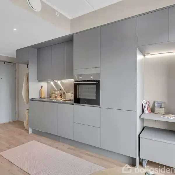 Grå køkkenenhed med integreret ovn og kogeplade langs en væg. Moderne og minimalistisk indretning med trælaminatgulv og en lille skrivebordsplads. Lysindfald fra spotlights på loftet.
