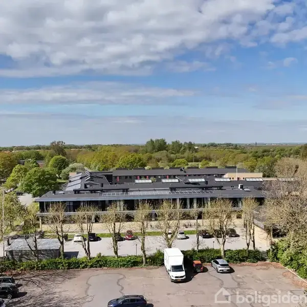 Et stort sort tag ligger over en række bygninger omkranset af træer. Foran ses parkerede biler, og i baggrunden strækker sig en grøn skov under en delvist skyet himmel.