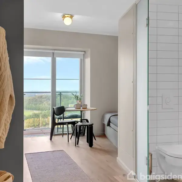 Dør ind til et lyst værelse med trægulv; bord og to stole ved vindue med udsigt. Badeværelse med glasdør til højre. Strikket trøje hænger på væggen.