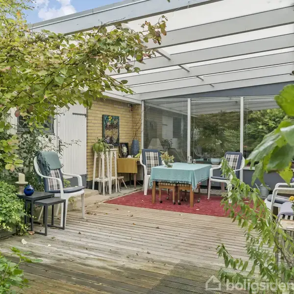 Terrassen med havemøbler står under en glastag i en frodig have. Der er et bord med blå dug omgivet af stole, og vegetation vokser tæt omkring.