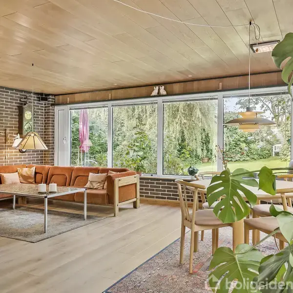 Brunt, åbent rum med orange sofagruppe foran store vinduer, der giver udsigt til frodig have. Rundt spisebord med stole, grønne planter og en stålampe skaber en hyggelig atmosfære.