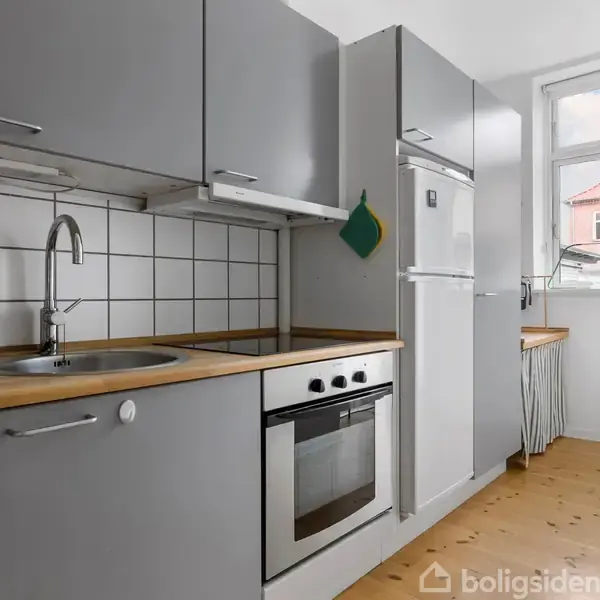 Grå køkkenskabe med indbygget ovn og køleskab placeres langs væggen. Træbordplader og hvidt flisebaggrund. Et vindue med lysindfald og en vase med blomster i hjørnet.