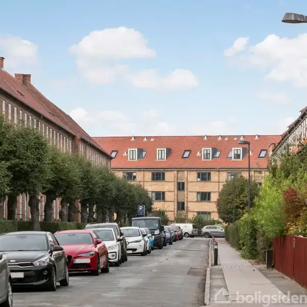 En række parkerede biler står langs en smal vej, flankeret af røde murstensbygninger med skifertage. Træer står i række langs fortovet under en blå himmel med skyer.