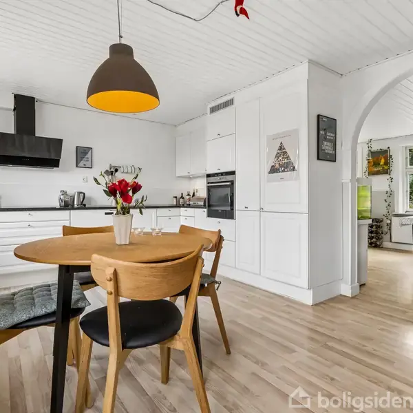 Rundt træbord med fire stole er centralt placeret, dekoreret med en vase med blomster. Bagved findes et moderne, hvidt køkken med hængende lamper, åbne hylder og store vinduer.