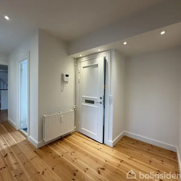 En entrédør står åben i en lys gang med en trægulv. På væggen hænger en telefon ved siden af en radiator. Spotlys oplyser rummet, og en trappe ses i baggrunden.