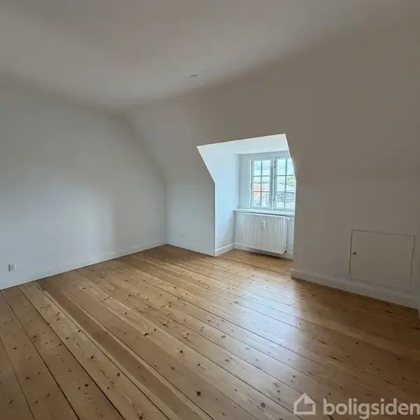 Et tomt rum med træplankegulv og hvide vægge. En indhak med et vindue og en radiator viser udenfor til bygninger. Rummet er lyst og minimalistisk.