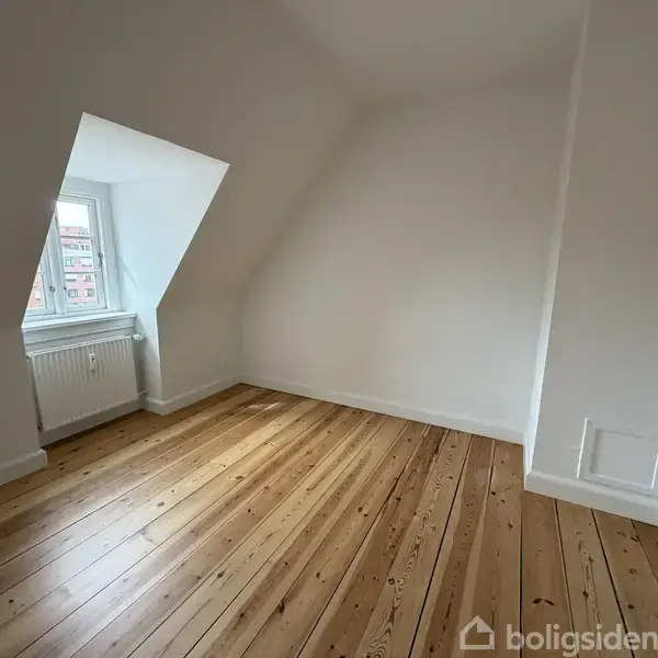 Et lyst værelse med et skråt loft har trægulv og et lille vindue i hjørnet, der giver udsigt til omkringliggende bygninger. Rummet er tomt med hvidmalede vægge.