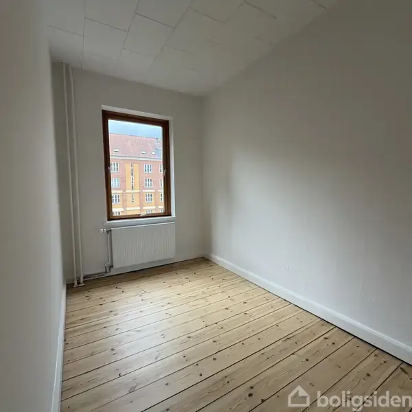Et lille, tomt værelse med trægulv og hvidmalede vægge. Et vindue lukker lys ind, og udenfor ses en rødstensbygning. En radiator er placeret under vinduet.