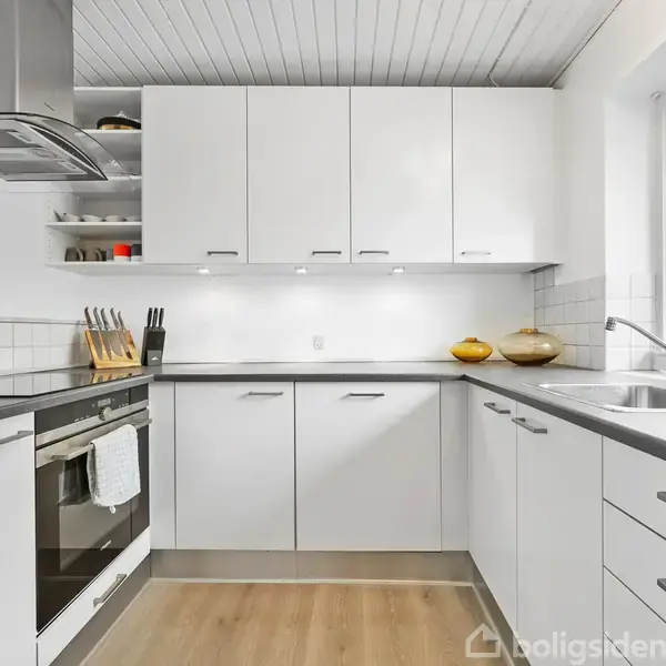 Et moderne køkken med hvide skabe og sorte bordplader. En ovn og komfur under emhætte; køkkenredskaber står ved siden af. Store vinduer giver udsigt til et udendørs område.