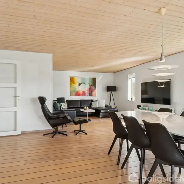 Stue med træloft og trægulv, indeholder spisebord med sorte stole. I hjørnet er en lænestol med fodskammel. På væggen hænger et farverigt maleri, og der er vinduer med udsigt.