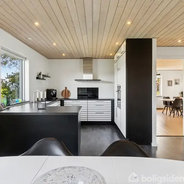 Et moderne køkken med sort-hvide elementer og integrerede apparater. Rummet har træloft med spotlys, store vinduer til venstre, og en åbning til en tilstødende spisestue.