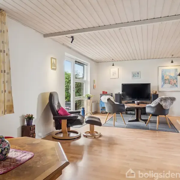 Stue med trægulv og hvide vægge, indeholdende en grå sofa, lænestole, fladskærmsfjernsyn på et skab, og spisebord med stole. Store vinduer slipper lys ind i rummet.