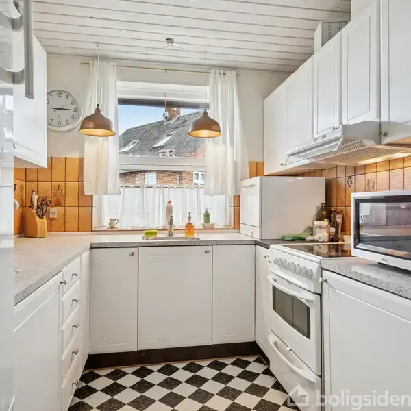 Et hvidt køkken med ovn, mikroovn og køleskab i brug. To hængende lamper og vindue med udsigt til bygninger udenfor; skakternet gulv og brune fliser på væggen.