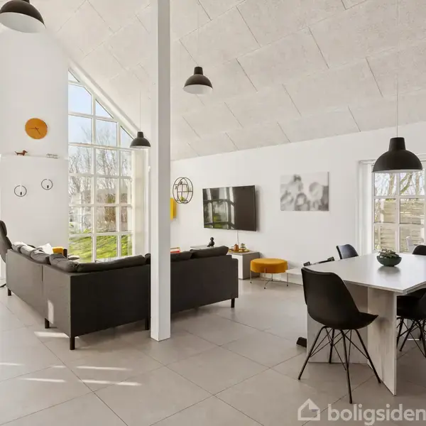 Stor, lys stue med højt loft indeholder en sort sofa, spisebord med stole, vægmonteret TV. Store vinduer giver udsigt til grøn have. Indretningen er moderne med sorte og orange elementer.