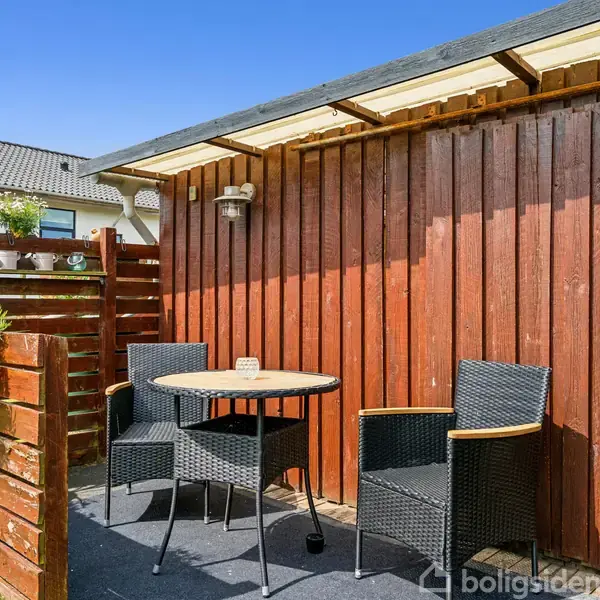 To flettede stole og et rundt metalbord står på en terrasse foran en rød trævæg. Omkring er det blå himmel, græs og blomster i potter på en lav træhegn.