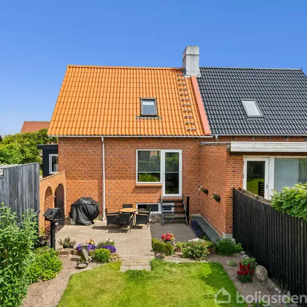 Rødt murstenshus med orange og sort tag står i en velplejet have med terrasse møbler og grill omgivet af høj sort træhegn og blomsterbede under en klar blå himmel.