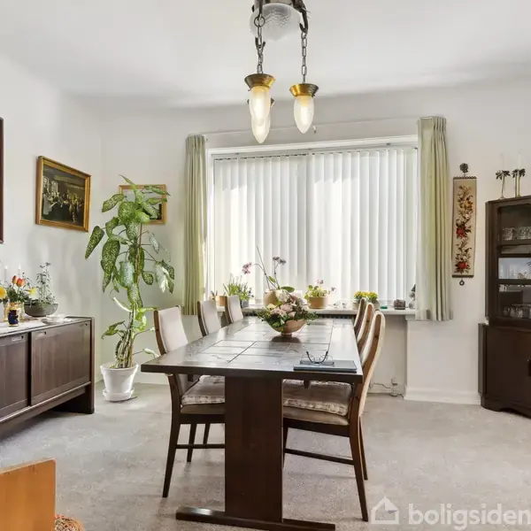 Et spisestuebord med seks stole står i et lyst rum. Over bordet hænger en lysekrone. Rummet er dekoreret med planter, malerier, en kommode og en vitrineskab ved vinduet.