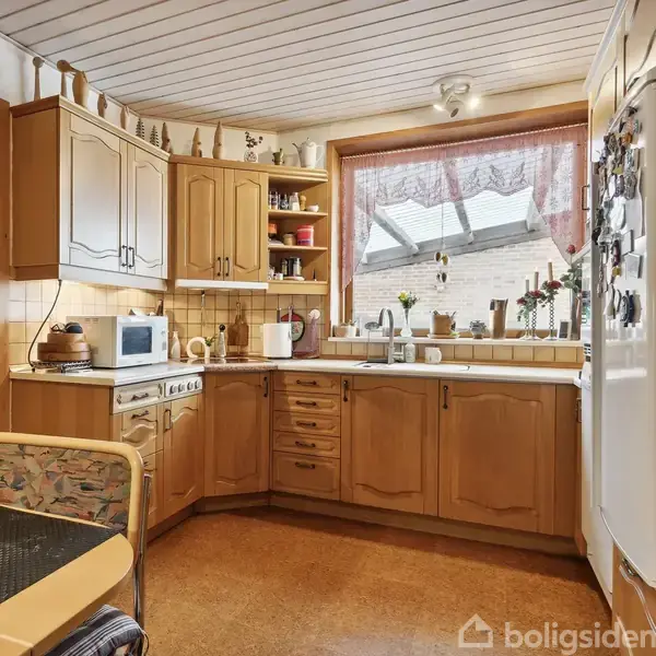 Køkken med træskabe omgiver en kogeplade og mikroovn. En stor vindueskarm med planter og lys filtrerer sollys. En stol står ved et bord.