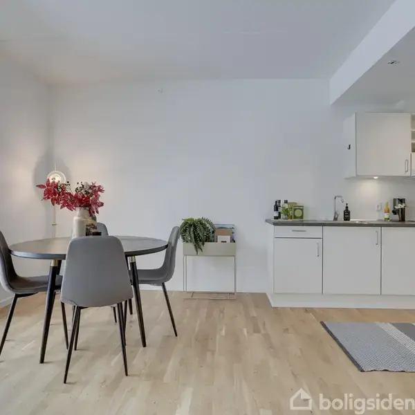 Rundt bord med tre stole står i et lyst rum. Der er en bordlampe med røde blomster ved siden af. Bagved ses et hvidt køkken med moderne apparater.