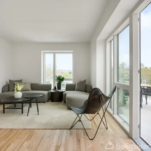Lys stue med en grå hjørnesofa og stole placeret omkring runde borde. Store vinduer åbner op til en balkon med udsigt over omgivelserne. Væggene er hvide.