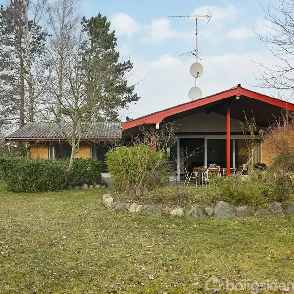 Et enfamiliehus med rødt tag og flere satellit-skåle står i en have med grønne buske og træer. Der er havemøbler på en terrasse foran huset.