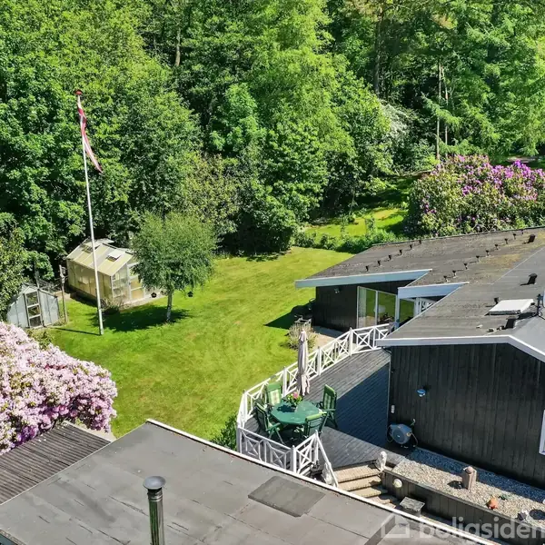 Et træhus med sort tag ligger i en frodig have. En terrasse med siddepladser strækker sig ud, mens en flagstang og blomstrende buske omkranser græsplænen i naturen.
