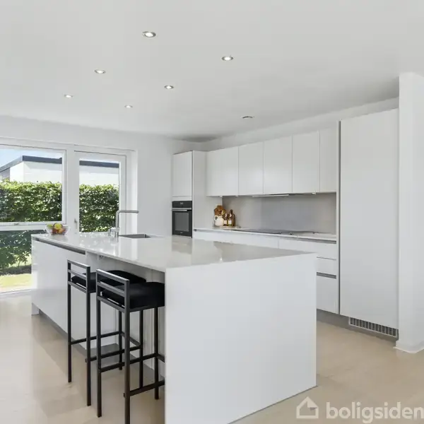 En L-formet moderne villa med lys murstensfacade og sorte tagsten står på en velholdt græsplæne under en klar blå himmel, omgivet af lave hække.