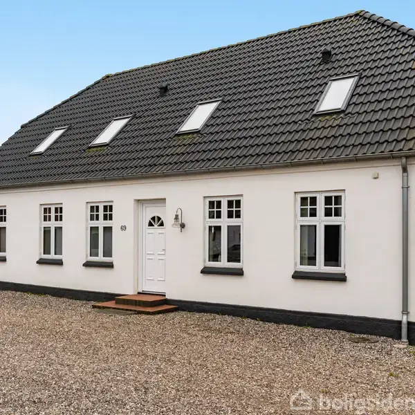 Et hvidt hus med sort tegltag står stille. Det har flere vinduer og en hvid dør. Gruset indkørsel og græsplæne omgiver det i et roligt område.