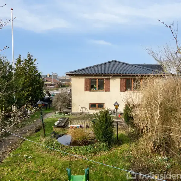 Hus med fladt tag står i en have omringet af træer og buske. En træterrasse og en flagstang ses til venstre. Himlen er klar.