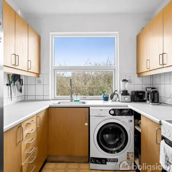 Køkken med hvidt vaskemaskine integreret i bordpladen, omgivet af træskabe og flisebelagte vægge. Et vindue giver udsyn til naturen udenfor. Magnetbogstaver på køleskabet.