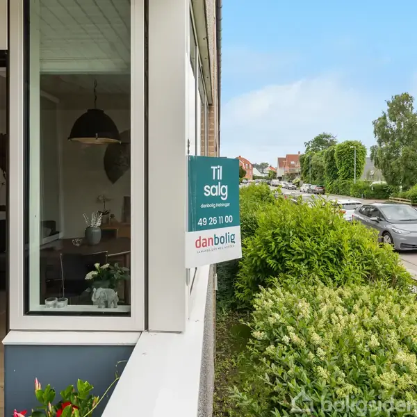Bolig til salg skilt hænger på murstensbygning ved siden af en stor busk. Tekst på skiltet: "Til salg", "danbolig", "49 26 10 00". Indendørs ses en verdenskort plakat.