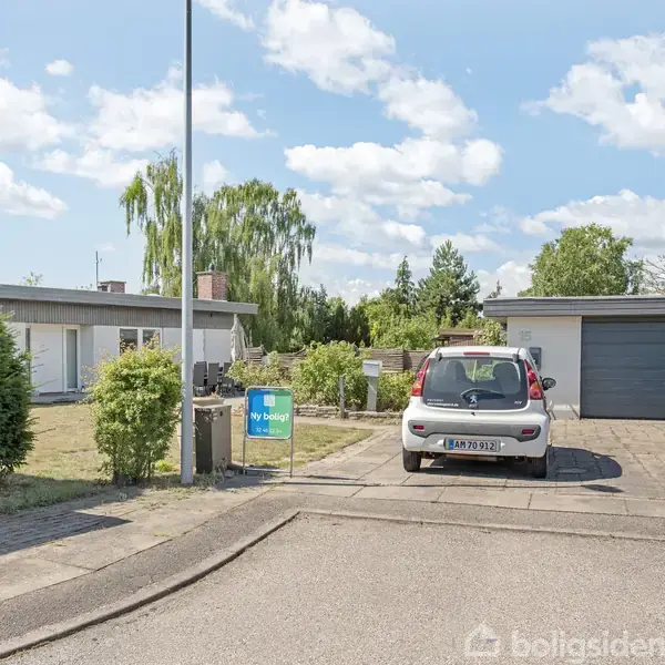 En bil holder parkeret foran en garage i en indkørsel. Der er en trailer ved siden af bilen. Baggrunden består af et hvidt hus omgivet af grønne buske og træer. Et skilt ved vejen siger "Til salg".