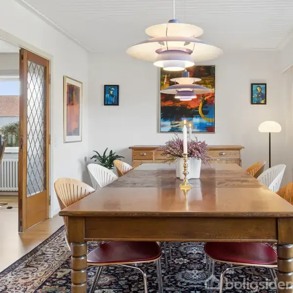 En spisebord med to tændte lysestager står i et rum med en træstol. Baggrunden har et kunstbillede på en hvid væg, set gennem en glasdør med træramme.