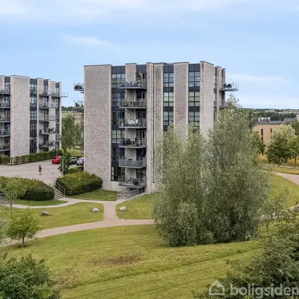 En moderne, fleretagers bygning står roligt omgivet af grønne græsarealer og træer, med flere stier, der krydser gennem området. Andre lignende bygninger ses i baggrunden.