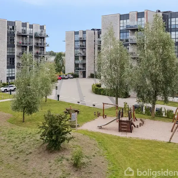 Moderne lejlighedskomplekser står stille i baggrunden med store vinduer. I forgrunden er der en legeplads med gynger og træklatringtårn omgivet af grønne græsplæner og træer.