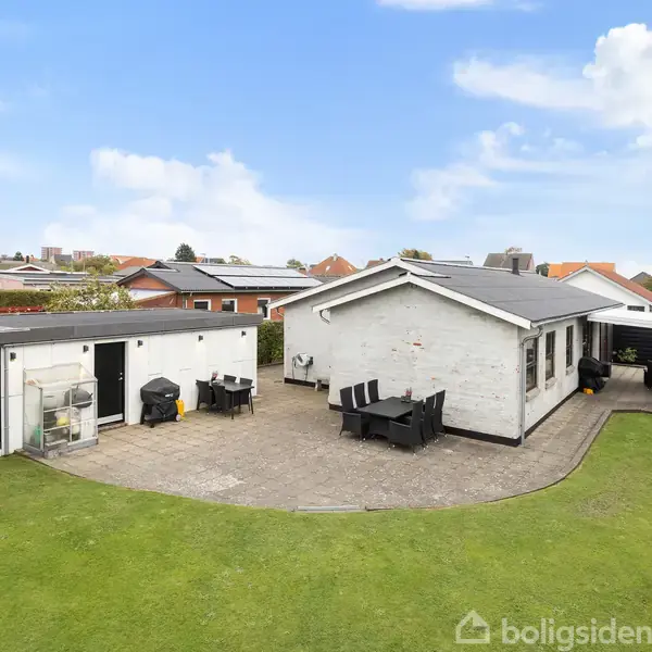 Huset står stille i en forstadshave med grøn græsplæne og flisebelagt terrasse. Omgivet af tætte buske og andre huse under en blå himmel med hvide skyer.