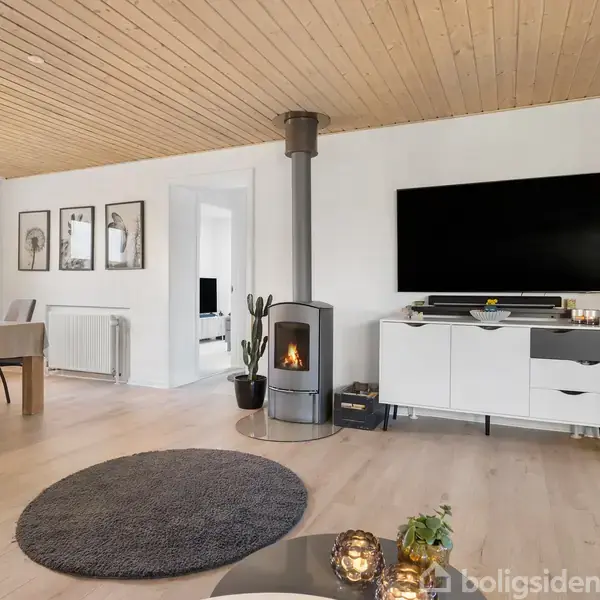 En stue med et moderne tv på væggen, under en brændeovn i hjørnet og et rundt tæppe på gulvet. Til venstre står et spisebord med stole og hængelamper.