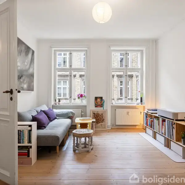 Stue med en grå sofa fyldt med farverige puder, et sofabord, og en bogreol langs en væg. Vinduerne viser fjerntliggende bygninger. Rummet er lyst og moderne indrettet.